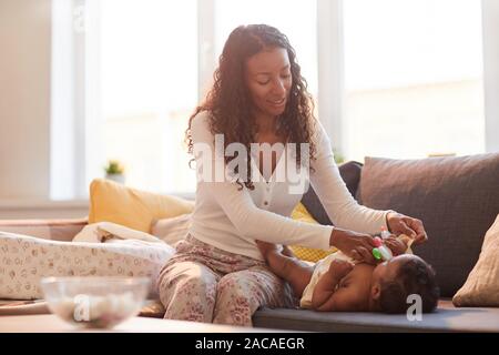 Portrait d'entraide aux teintes chaleureuses mère afro-américaine s'habiller son bébé sur la table contre la lumière du soleil, copy space Banque D'Images