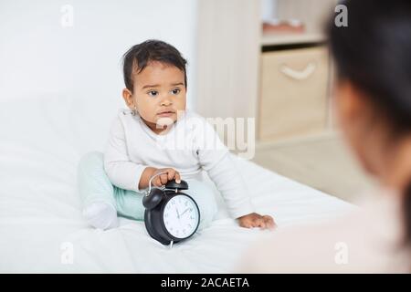 Petit enfant assis sur le lit et jouer avec réveil tout en regardant la mère Banque D'Images