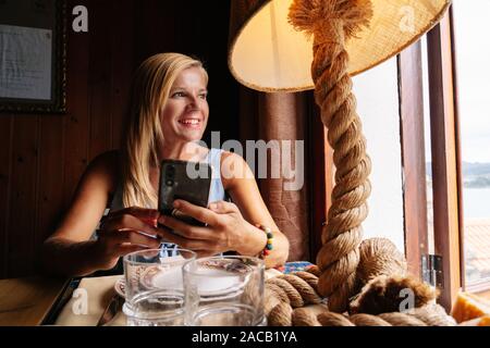 Une belle femme blonde caucasienne utilise le téléphone mobile dans un restaurant à côté d'une fenêtre Banque D'Images