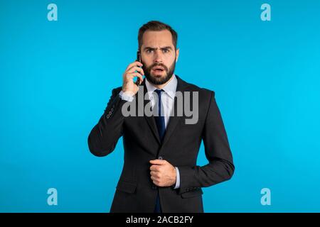 Businessman parle au téléphone, entendu de mauvaises nouvelles et exprime sa stupéfaction, choc isolé sur fond studio bleu. Portrait de bel homme. Banque D'Images