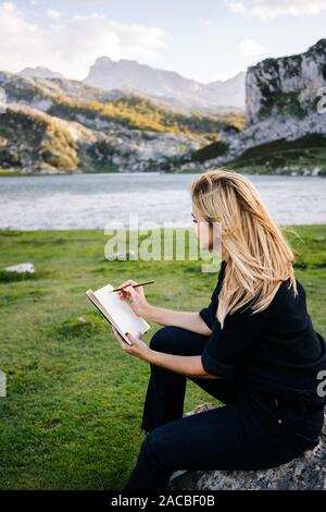 Une belle femme blonde caucasienne écrit et s'appuie sur un bloc-notes dans un paysage de montagne avec lac Banque D'Images