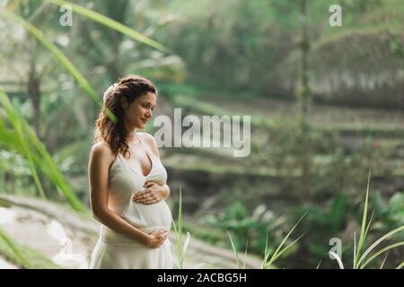 Jeune femme enceinte en robe blanche avec vue sur les rizières en terrasses de Bali dans la lumière du soleil du matin. Harmonie avec la nature. Concept de la grossesse. Banque D'Images