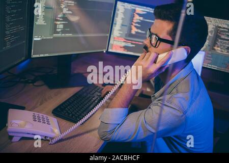 Photo du profil de l'informaticien guy travailler tard dans la nuit assis à la chaise de bureau téléphone parlant moniteurs experts débogage site web service correct Banque D'Images