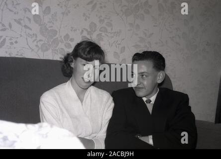 Années 1950, historique, un couple adolescent assis ensemble sur un canapé, garçon portant une veste, chemise et cravate, jetant un coup d'œil sur la fille, qui regarde directement la caméra, Angleterre, Royaume-Uni Banque D'Images