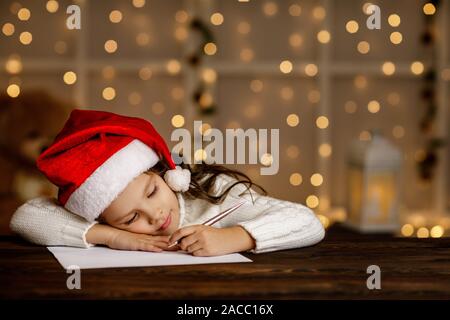 Heureux petit enfant girl in santa hat écrit la liste de souhaits ou une lettre au Père Noël sur fond de lumières. Joyeux Noël Banque D'Images