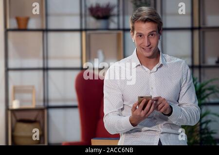 Jeune homme souriant dans un casque sans fil, portrait à l'intérieur loft, chambre élégante. Un gars positif utilise un smartphone pour la formation en ligne. Banque D'Images