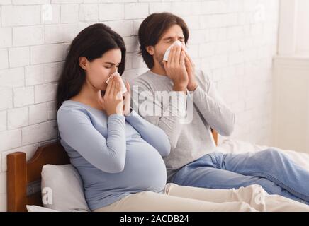 Pregnant couple se sentir malade, assis sur le lit et soufflant de nez Banque D'Images