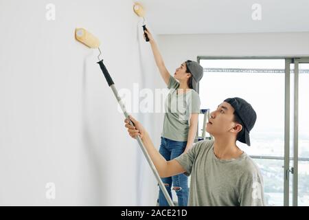 Les jeunes peintres professionnels asiatiques en majuscules à l'aide de rouleaux spéciaux lors de la peinture blanche des murs chambre Banque D'Images