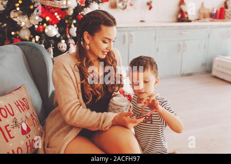 Jeune mère avec son fils dans la nouvelle cuisine années près de l'arbre de Noël cadeaux choisit au téléphone, elle est titulaire d'une tasse de café ou de thé. Banque D'Images