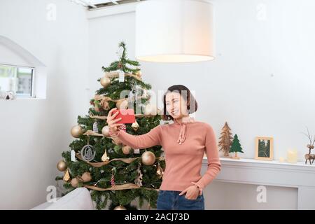 Très jolie femme en tenant à la maison tout en selfies debout près de l'arbre de Noël Banque D'Images
