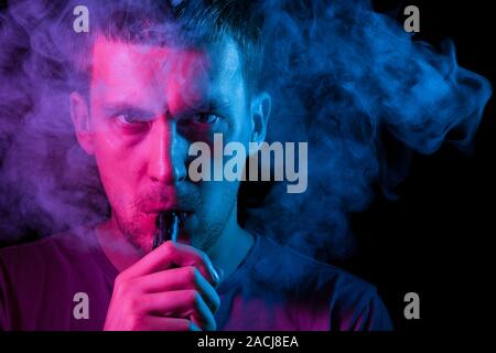 Un homme tient dans sa main et tire sur une cigarette électronique avec un vape en fumant et rejets de fumée à l'autre, son visage est mis en évidence avec b Banque D'Images
