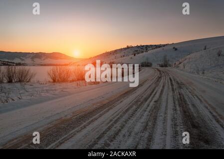 Paysage pittoresque avec l'asphalte sinueuse route enneigée par un col dans les montagnes couvertes de neige au coucher du soleil. La Russie, de l'Altaï Banque D'Images