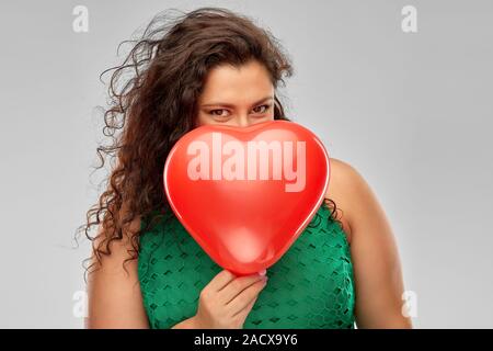 Valentine's Day, les gens et l'amour concept - femme en robe verte ludique se cacher derrière red heart shaped balloon sur fond gris Banque D'Images