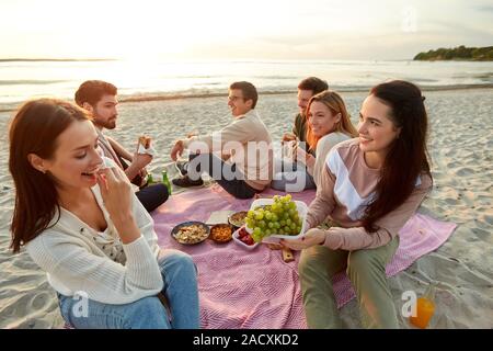 Amitié, loisirs et de fast food concept - groupe d'amis heureux de manger des sandwichs ou des hamburgers à pique-nique sur la plage en été Banque D'Images