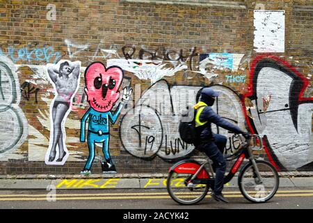 Un cycliste passant un graffiti sur un mur de brique à Buxton Street, près de Brick Lane Whitechapel London England UK Banque D'Images