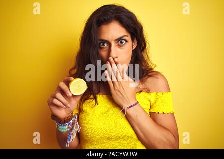 Belle Jeune femme tenant debout au-dessus de citron au fond jaune isolé couvrir la bouche à part choqué avec honte pour l'erreur, d'expression o Banque D'Images