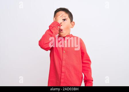 Bel enfant boy wearing red shirt élégant debout sur fond blanc isolé avec des pics de shock couvrant le visage et les yeux avec la main, regardant à travers Banque D'Images