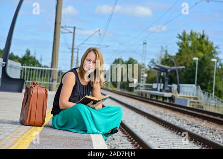 Jeune femme séduisante en longue jupe assise sur la gare et la lecture d'un livre Banque D'Images