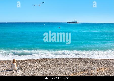 Une femme assise sur la plage de Menton, France sur la côte d'Azur comme un petit bateau et bateau plus grand cross à l'avant et une mouette vole au-dessus de Banque D'Images