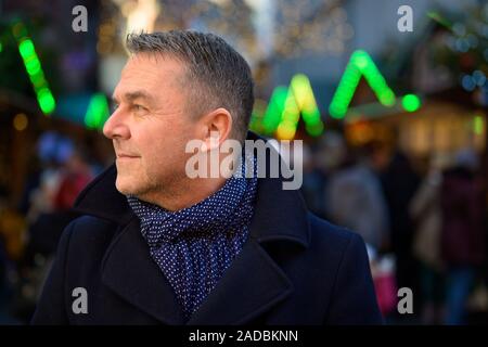 Homme d'âge moyen attrayant en hiver à la mode à la tenue de côté en regardant l'extérieur dans une place urbaine sur un soir de fête Banque D'Images