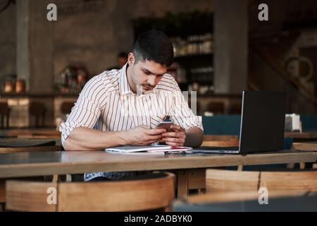 La lecture de certaines informations d'un smartphone. Homme adulte est assis dans un café au jour et à l'aide de l'ordinateur portable pour le travail à distance Banque D'Images