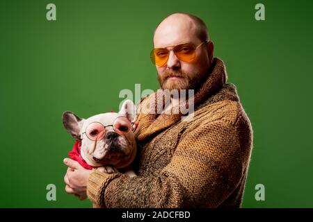 Homme sérieux dans les verres avec bouledogue français dans ses bras sur fond vert vide Banque D'Images