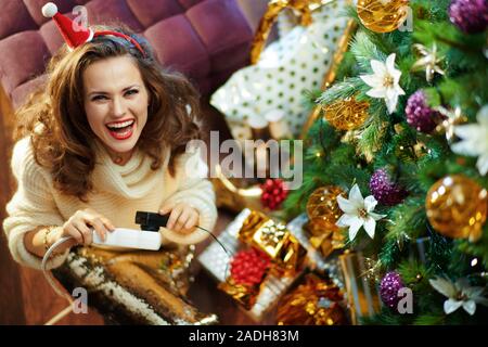 Happy housewife avec de longs cheveux de brunette en or paillette jupe et pull blanc sous l'arbre de Noël décoré des boîtes près de l'actuelle de l'insertion de câble Banque D'Images