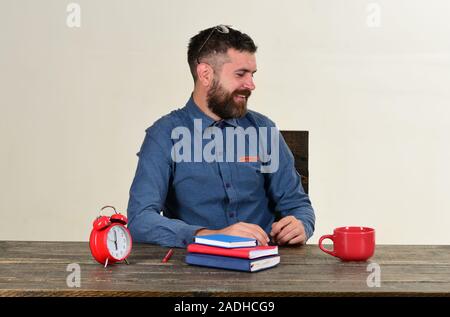 Homme à barbe est titulaire d'ordinateur isolé sur fond blanc. Examen et étude de concept. Avec des enseignants professionnels visage souriant est assis à un bureau en bois. Tasse et rétro avec horloge rouge et bleu livres sur table vintage Banque D'Images
