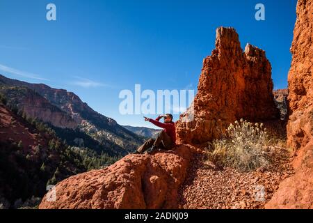 Un male hiker est situé au sommet d'une formation de roche rouge qui rappelle le Parc National de Bryce Canyon. Il montre au loin dans quelque chose. Banque D'Images