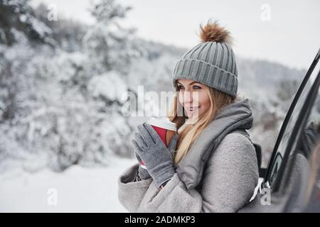 Pour vous réchauffer, boissons fraîches. Jolie fille dans des vêtements chauds debout dans l'hiver tout en bois s'appuie sur la voiture et la tenue de tasse de café Banque D'Images