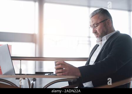Patron est assis sur la chaise près de la table de couleur argent avec ordinateur portable et est titulaire d'tasse de café Banque D'Images