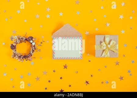 Vue supérieure de la composition festive avec couronne de fête, carte de voeux vierge avec enveloppe et boîte-cadeau décoration lumineuse sur fond jaune avec sparkle st Banque D'Images