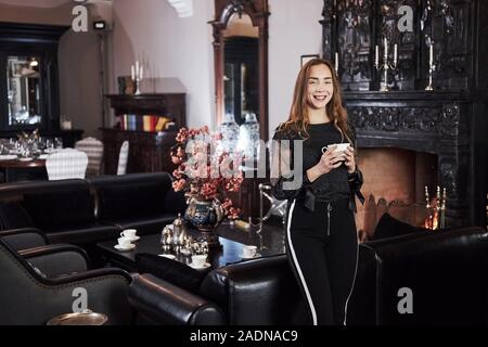 Portrait of smiling girl avec blanc tasse de thé dans les mains et se dresse contre cheminée Banque D'Images