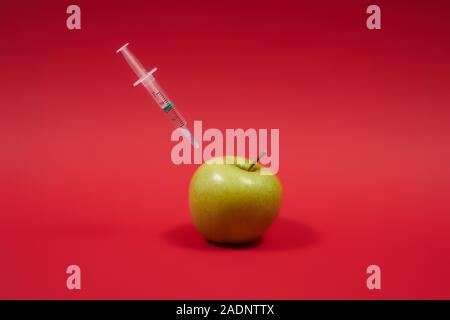 Le poison pénètre dans la pomme verte. Seringue avec liquide dans les fruits. Photo concept OGM. Nourriture empoisonnée sur le fond rouge. Banque D'Images