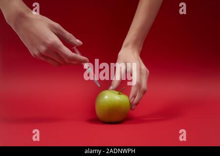 Les belles mains de la femme injectent du poison dans la pomme verte. Seringue avec liquide dans les fruits. Photo concept OGM. Nourriture empoisonnée sur le fond rouge. Banque D'Images