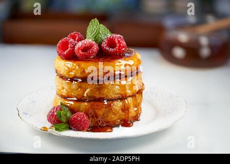 Vue de face de trois crêpes au fromage avec du miel et garni de framboises, le sucre et les feuilles de menthe sur un arrière-plan flou. Banque D'Images