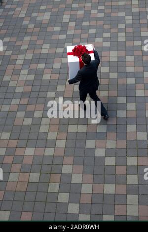 Un jeune homme portant un grand cadeau blanche entourée d'un arc rouge en flânant dans une cour Banque D'Images
