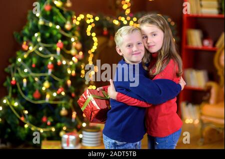 Donner des cadeaux enfants heureux tout en se tenant dans une chambre sur l'arrière-plan d'un arbre de fête. Hug et sourire. Noël et Nouvel An Banque D'Images