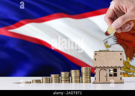 Man holding coins mise en maison en bois tirelire, de brandir le drapeau des Samoa américaines dans l'arrière-plan. Économiser de l'argent pour prêt hypothécaire. Banque D'Images