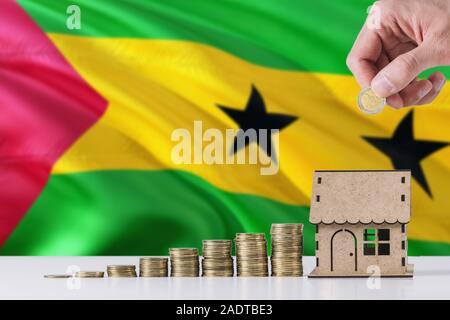 Man holding coins mise en maison en bois tirelire, Sao Tomé-et-Principe drapeaux à l'arrière-plan. Économiser de l'argent pour prêt hypothécaire. Banque D'Images