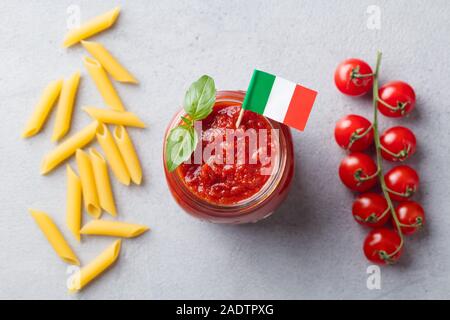 Sauce tomate traditionnelle dans un bocal en verre avec drapeau italien, pâtes penne sur un côté. Fond gris. Vue d'en haut Banque D'Images