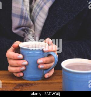 Deux tasses bleu de cacao sur une table en bois, dont un qui est tenu dans les mains d'une femme dans un manteau chaud. Nouvelle couleur à la mode. Banque D'Images