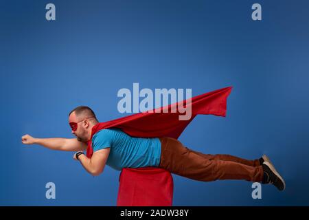 Homme en rouge cape de super-héros volant sur fond bleu vide en studio Banque D'Images