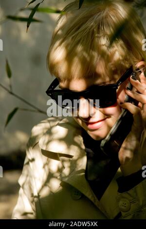 Jeune femme élégante, 20's style des années 1950, le port de lunettes de soleil, imperméable et vu par le feuillage à l'aide de téléphone mobile Banque D'Images