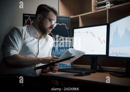 Certains documents. Homme barbu en chemise blanche travaille au bureau avec plusieurs écrans d'ordinateur dans les tableaux d'index Banque D'Images