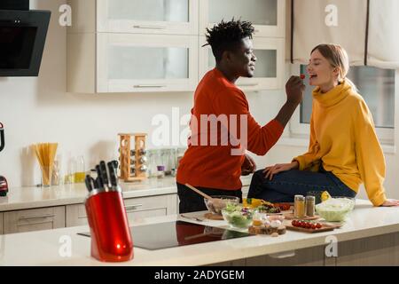 Genre jeune homme nourrir sa petite amie avec tomate cerise dans la cuisine Banque D'Images