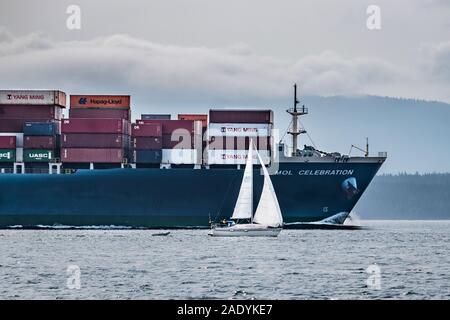 Deux personnes un petit yacht à voile à regarder en haut, un énorme navire conteneur chargé comme il dépasse, avec sa proue juste en avant du voilier. Banque D'Images