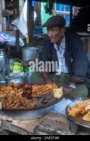 Un vieux homme birman dans un capuchon vend divers aliments frits d'un décrochage dans un marché de village à Kanne, nord-ouest de Myanmar (Birmanie) par la rivière Chindwin Banque D'Images