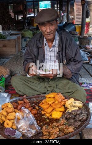 Un vieux homme birman dans un capuchon vend divers aliments frits d'un décrochage dans un marché de village à Kanne, nord-ouest de Myanmar (Birmanie) par la rivière Chindwin Banque D'Images