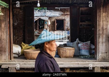 Une femme plus âgée dans un chapeau de riz conique passe par un milieu rural dans un entrepôt composé sur un village-rue dans le nord-ouest rural Myanmar (Birmanie) Banque D'Images